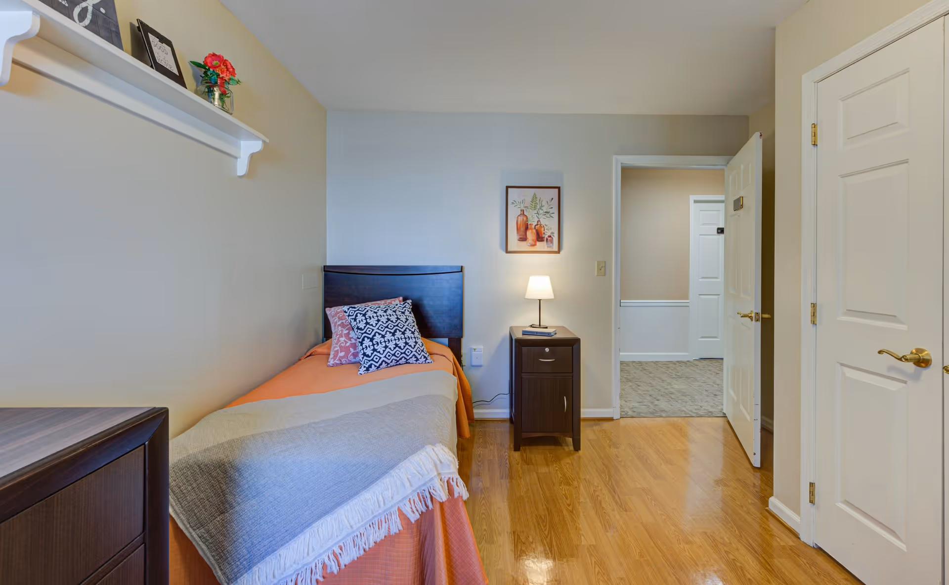 A small, neatly arranged bedroom with a single bed covered in an orange bedspread and a gray throw blanket. There are two decorative pillows on the bed. Next to the bed is a dark wooden nightstand with a small lamp and a framed picture hanging on the wall above it. The room has light-colored walls, hardwood flooring, and a white door that is partially open, showing a glimpse of a carpeted hallway outside. A white shelf with decorative items is mounted on the left wall.