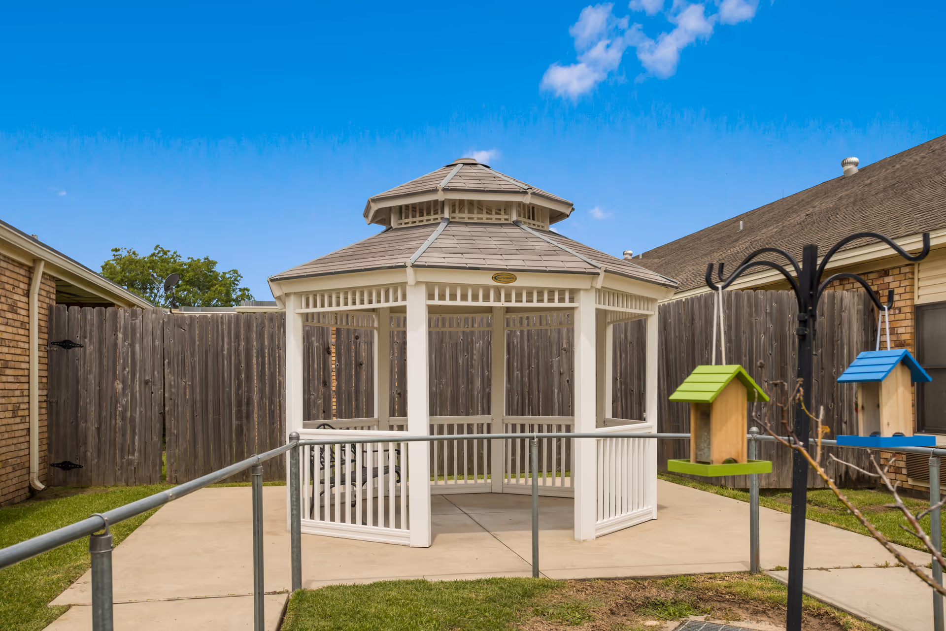 White wooden gazebo on a concrete pad in a fenced courtyard with birdhouses and a clear blue sky.