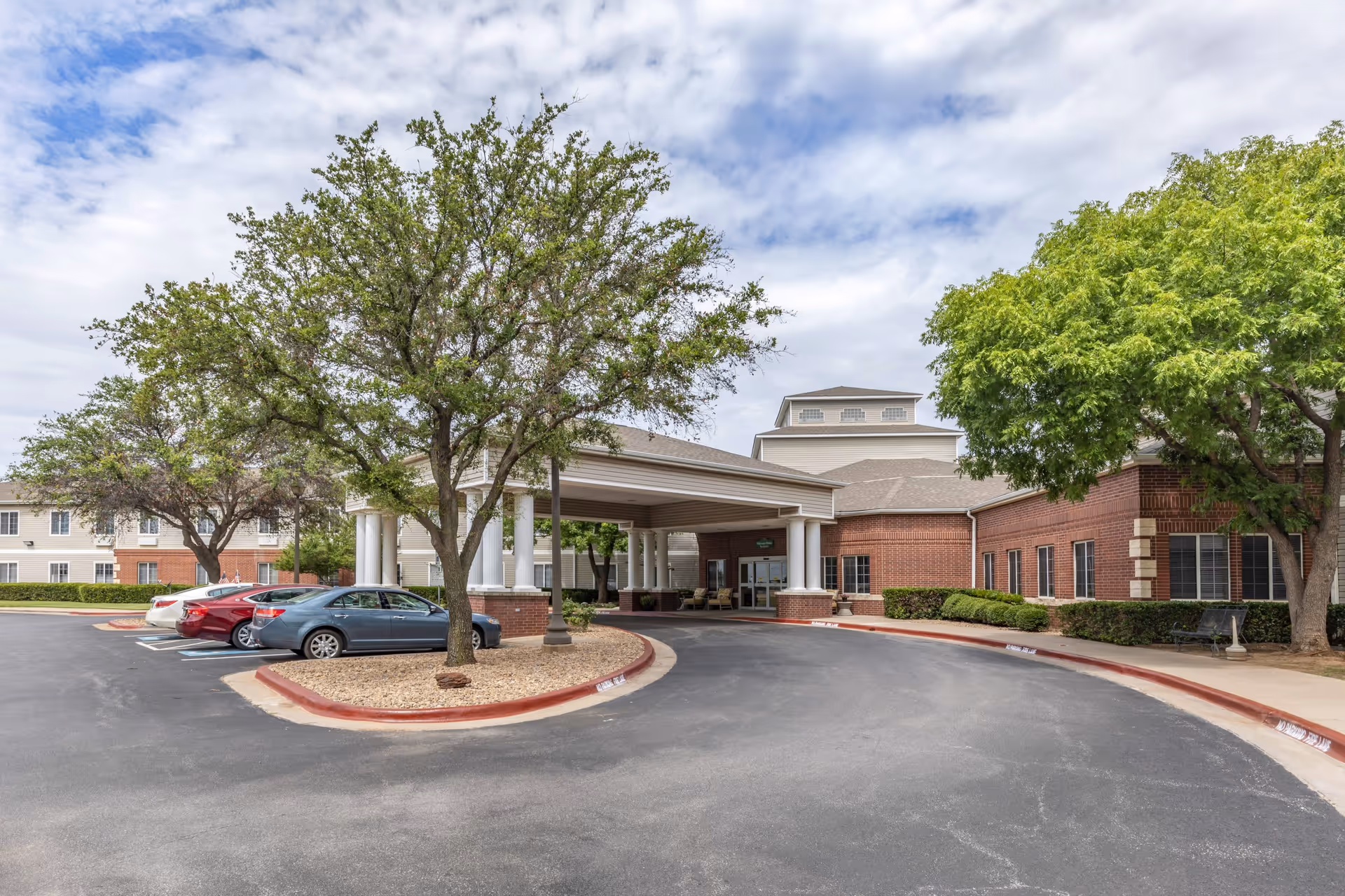 Exterior view of the entrance to Royal Estates of Wichita Falls, showing a covered drop-off area with white columns, a curved driveway, several parked cars, and trees with green foliage under a partly cloudy sky.