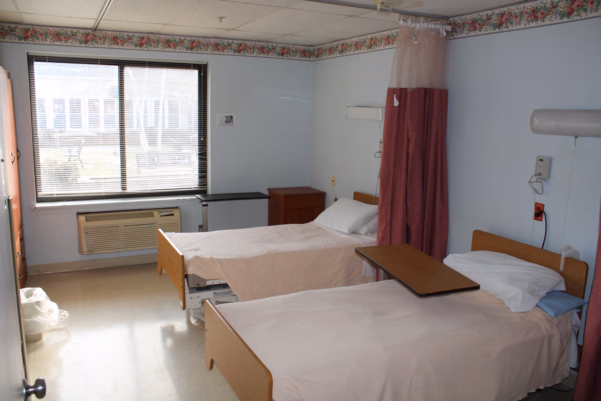 Two hospital-style beds in a shared care room with an overbed table, privacy curtain, window with blinds, and bedside cabinets.