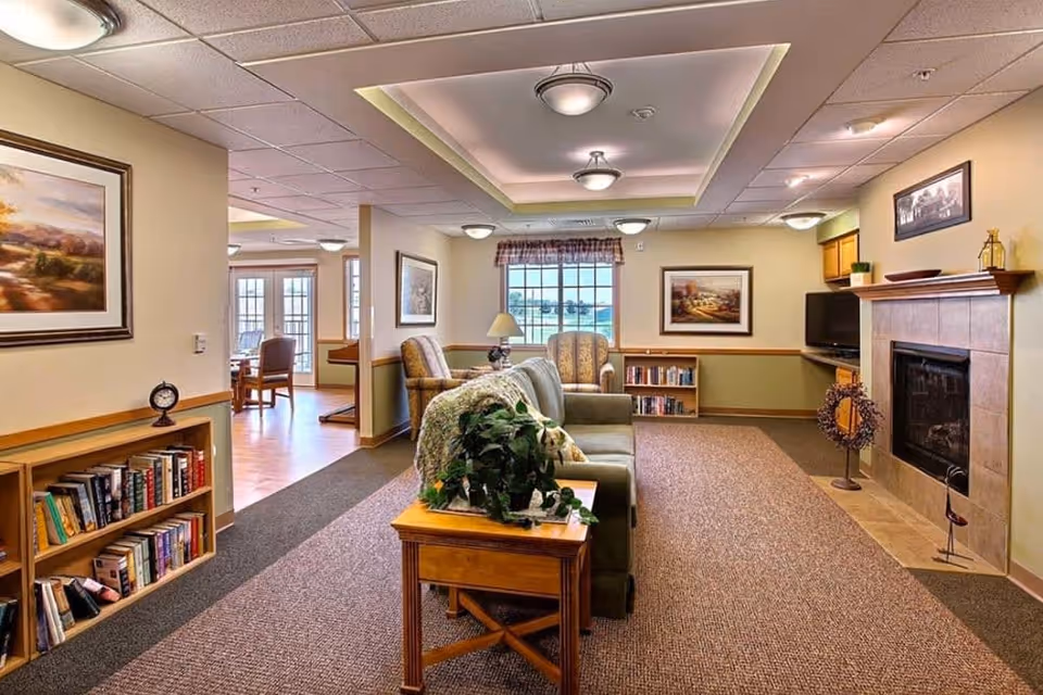 A bright, furnished common living area with couches and armchairs centered around a fireplace, bookshelves, and a window.
