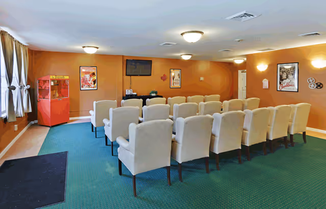 A small theater room with three rows of beige upholstered chairs facing a wall-mounted flat screen TV. The room has orange walls decorated with vintage movie posters and a red popcorn machine in the corner. The floor is carpeted in green, and there are windows with curtains on the left side.