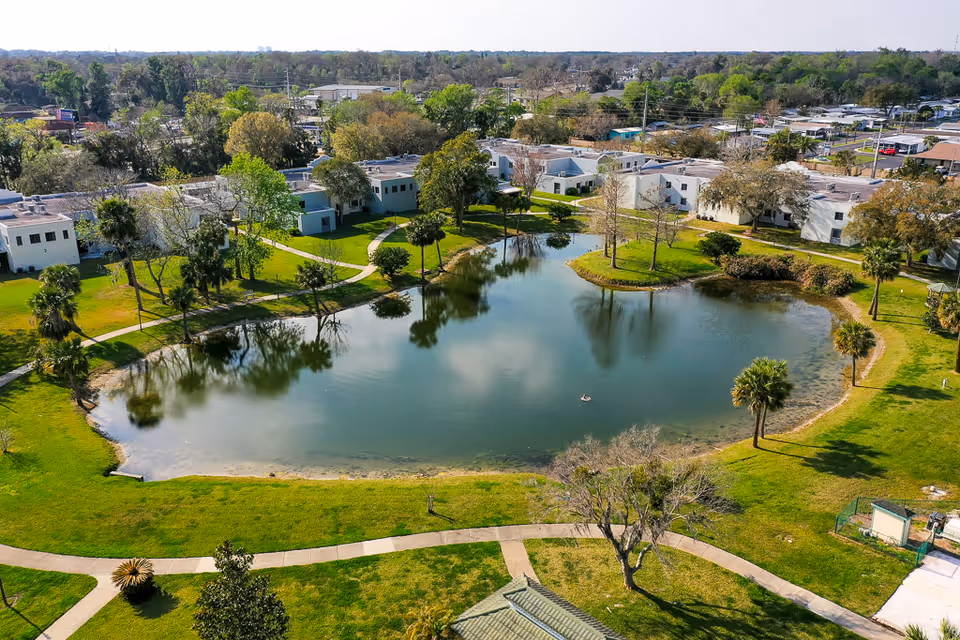 Aerial view of a senior living facility with a large pond surrounded by green grass, trees, and walking paths. Several white buildings are visible around the pond, with a residential neighborhood in the background under a clear sky.