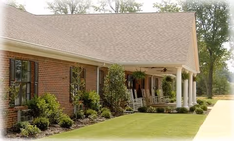 Exterior view of a brick building with a covered porch featuring white columns and several rocking chairs. The building is surrounded by neatly trimmed bushes and a well-maintained lawn.