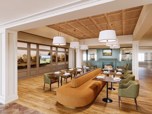 A spacious dining area in a senior living facility featuring a long, curved orange banquette with multiple dining tables set with plates and utensils. Green upholstered chairs surround the tables. The room has a wooden floor, a coffered ceiling with large white pendant lights, and a fireplace with a painting above it in the background. Large windows with scenic views line one wall.