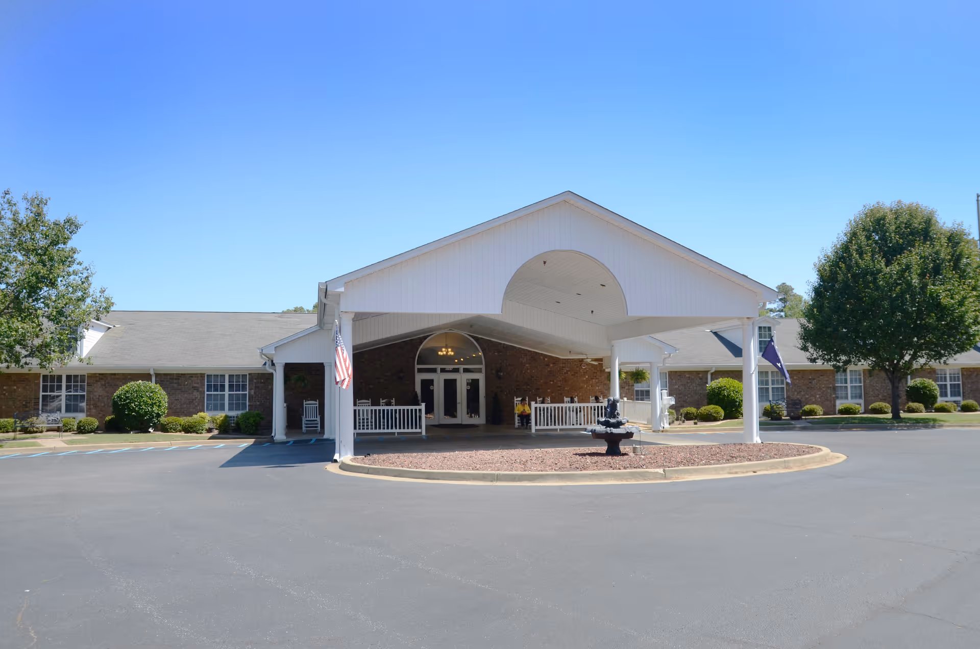 Front exterior view of The Gardens at Sumter facility with a covered entrance, a small fountain in the circular driveway, and landscaping including trees and bushes under a clear blue sky.