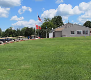 A large grassy lawn with a beige building in the background under a blue sky with scattered clouds. Three flagpoles with the American flag, a red flag, and a blue flag are visible near the building. Trees line the background behind the building.