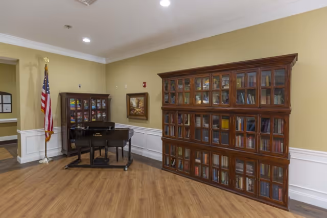 Interior room with wooden flooring featuring a black grand piano, two large wooden bookcases filled with books, an American flag on a stand, and a framed painting on the wall.