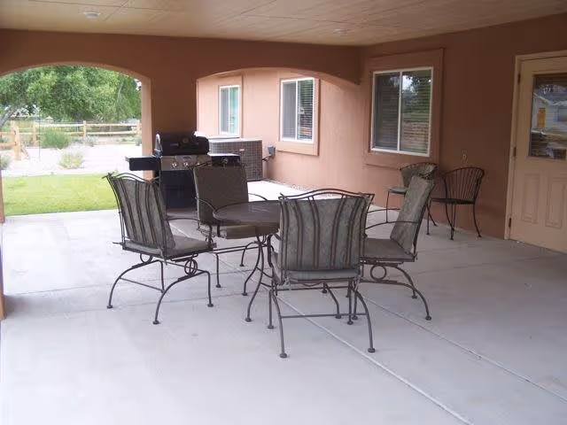 Covered outdoor patio area with a round metal table and four cushioned metal chairs. There is a barbecue grill in the corner near the wall of the building, and a grassy yard with a wooden fence is visible beyond the patio.