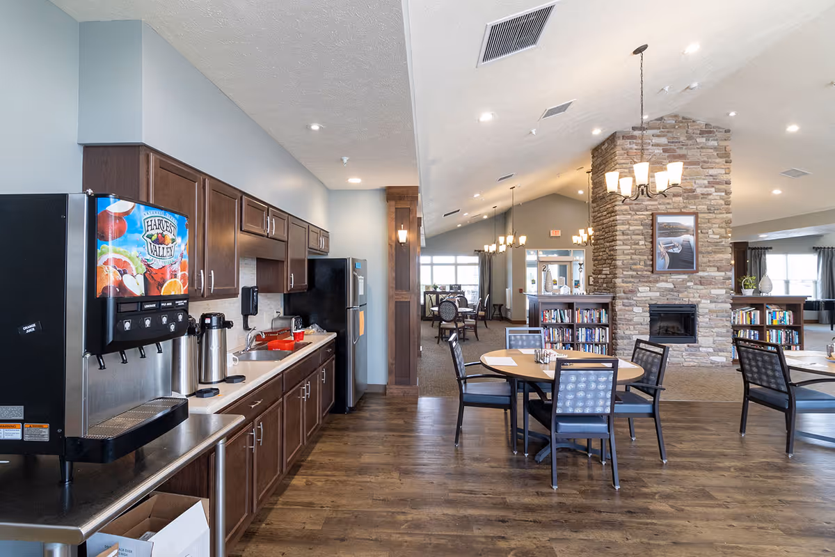 Interior view of a senior living facility common area with a kitchenette on the left featuring dark wood cabinets, a beverage dispenser, and a refrigerator. To the right, there are round tables with chairs, a stone fireplace with bookshelves on either side, and multiple hanging light fixtures. Large windows and a high ceiling create a bright and open atmosphere.