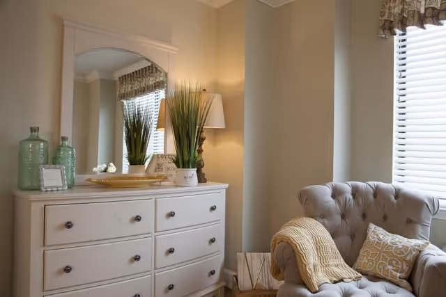 A cozy corner of a room featuring a white dresser with six drawers topped with decorative items including two green glass bottles, a small framed picture, a lamp, a potted plant, and a mirror. Next to the dresser is a tufted armchair with a yellow knitted throw blanket and a patterned cushion, positioned near a window with white blinds and a patterned valance.