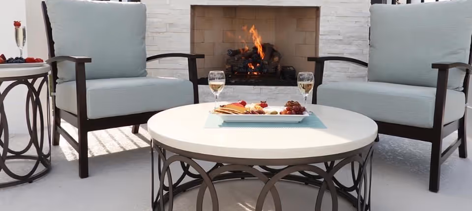 Two cushioned armchairs and a round coffee table with wine glasses and a snack plate in front of a lit fireplace.