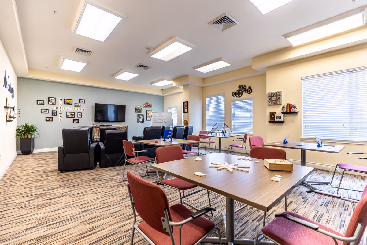 Bright communal activity room with tables and red chairs, a TV with recliners, and games and decor on the walls.