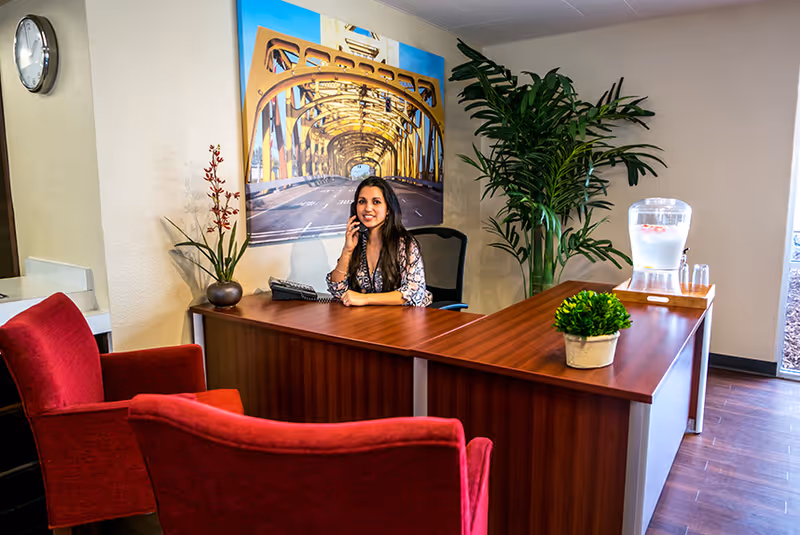 A woman sitting behind a wooden reception desk talking on the phone in a well-lit office area. The desk has a small potted plant and a water dispenser with glasses. There are two red upholstered chairs in front of the desk, a large green plant in the corner, and a framed picture of a yellow bridge on the wall behind her.