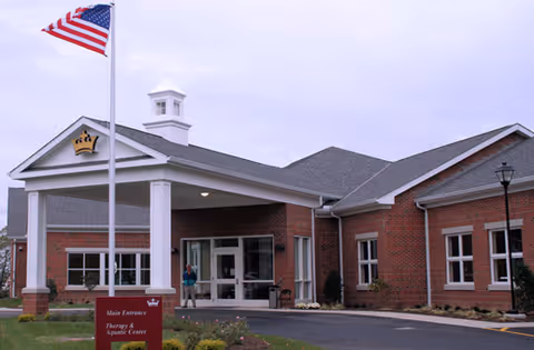 Exterior view of a single-story brick building with a covered entrance, an American flag on a flagpole in front, and a sign indicating the main entrance and therapy & aquatic center. The sky is overcast.