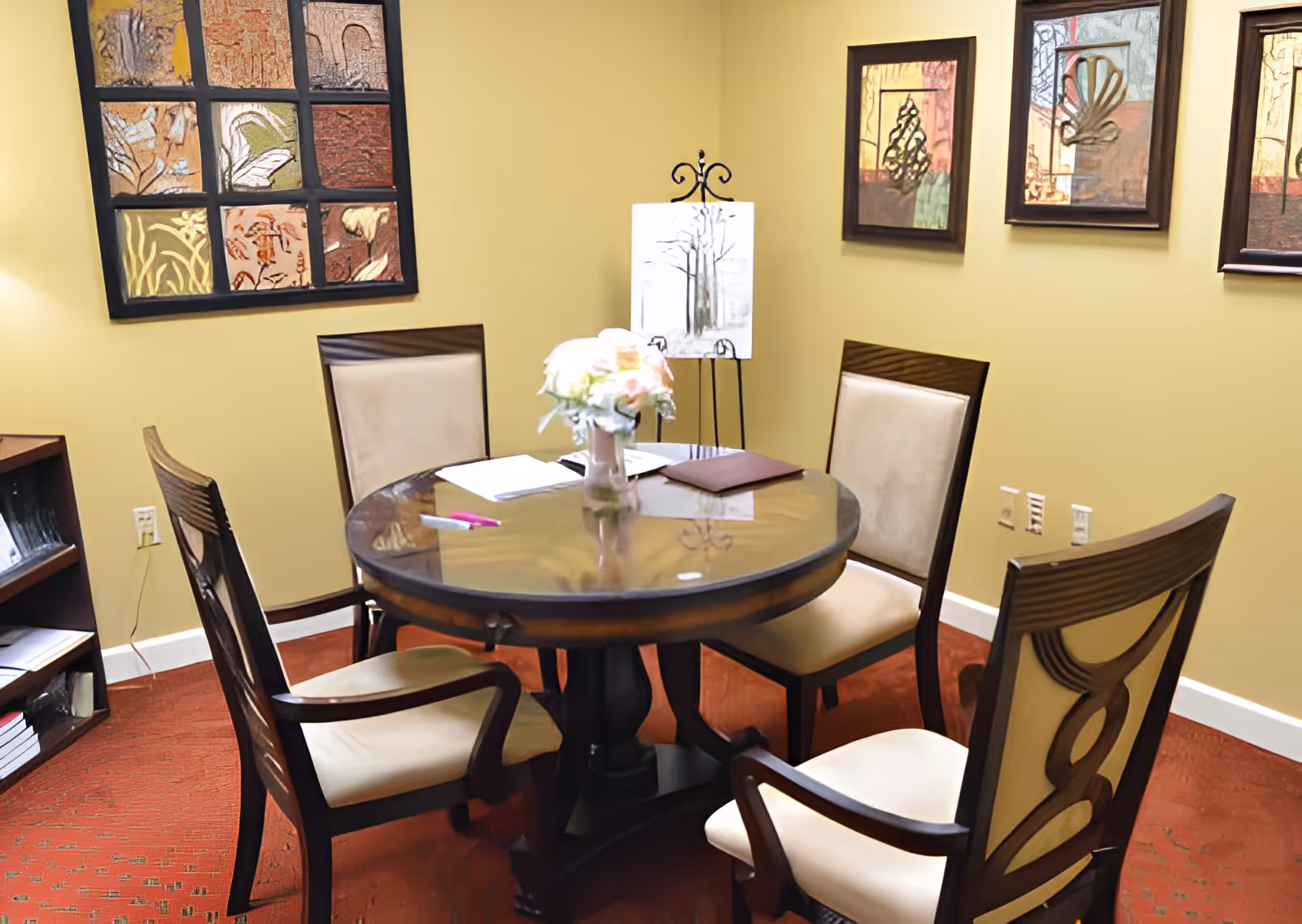 A small meeting or dining area with a round glass-top table surrounded by four cushioned wooden chairs. On the table, there is a vase with flowers, some papers, and a pen. The room has beige walls adorned with framed artwork and a small bookshelf in the corner.
