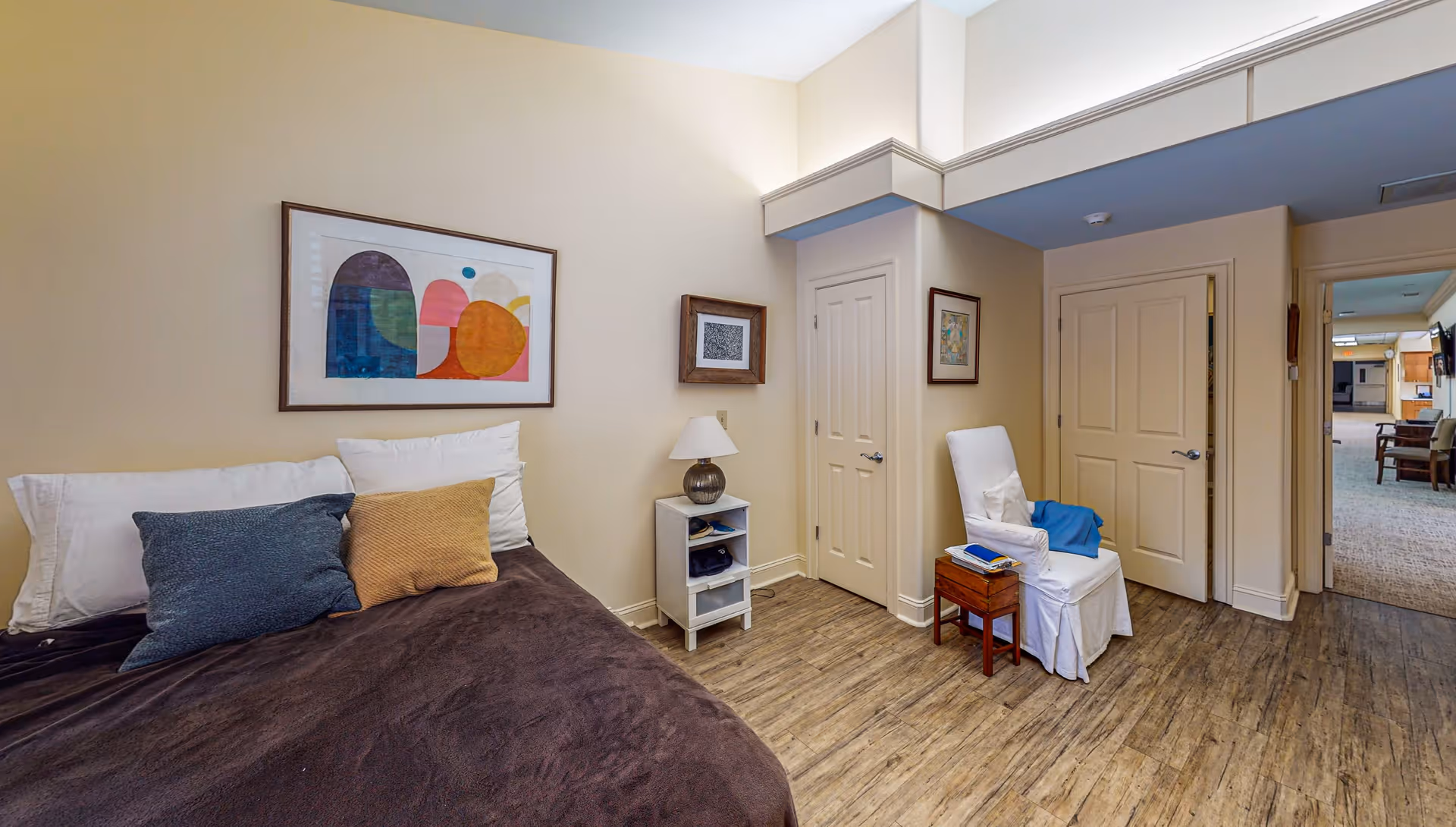 A cozy bedroom with a bed covered in a dark brown blanket and multiple pillows against a beige wall. Above the bed hangs a colorful abstract painting. Next to the bed is a small white nightstand with a lamp and some items on the shelves. A white armchair with a blue throw is positioned near two closed doors. The room has wood flooring and soft lighting, with a hallway visible in the background leading to another area with seating.