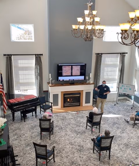 A spacious living room with high ceilings and large windows. There is a fireplace with a TV mounted above it, showing a presentation. Several chairs are arranged in a semi-circle facing the fireplace, with a few elderly people seated. A man wearing a mask stands near a whiteboard giving a presentation. The room has chandeliers, a piano, and an American flag near the windows.