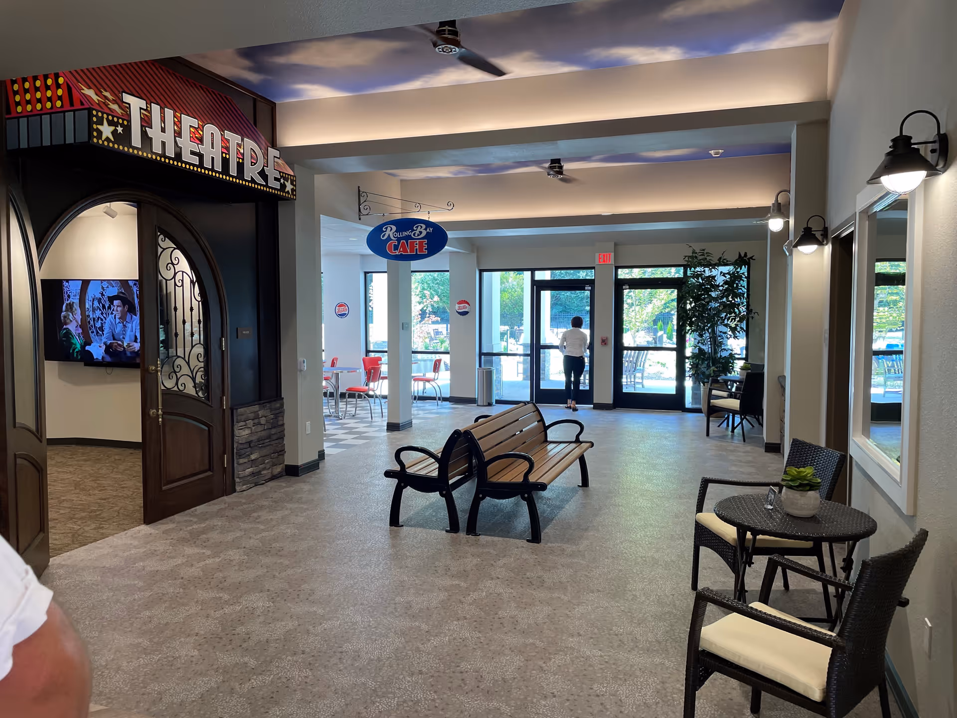 Interior view of a senior living facility showing an entrance to a theatre room with double wooden doors and a sign above reading 'THEATRE'. To the right, there is a small seating area with a round table, two chairs, and a potted plant. In the background, there is a cafe area with tables and red chairs, and a sign hanging from the ceiling that reads 'Rolling Bay CAFE'. A person is seen walking towards glass exit doors leading outside. The space is well-lit with ceiling lights and wall sconces.