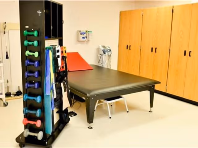 A physical therapy room with a black padded treatment table, a rack holding colorful dumbbells and resistance bands, wooden storage cabinets along the wall, and medical equipment mounted on the wall.