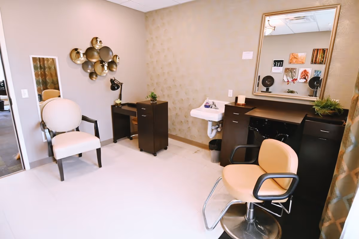 Interior of a salon area in a senior living facility featuring a beige salon chair with black armrests in front of a large mirror, a small white sink, dark wood cabinetry, a beige upholstered armchair, a decorative wall mirror with circular metallic discs, and a desk lamp on a small desk.