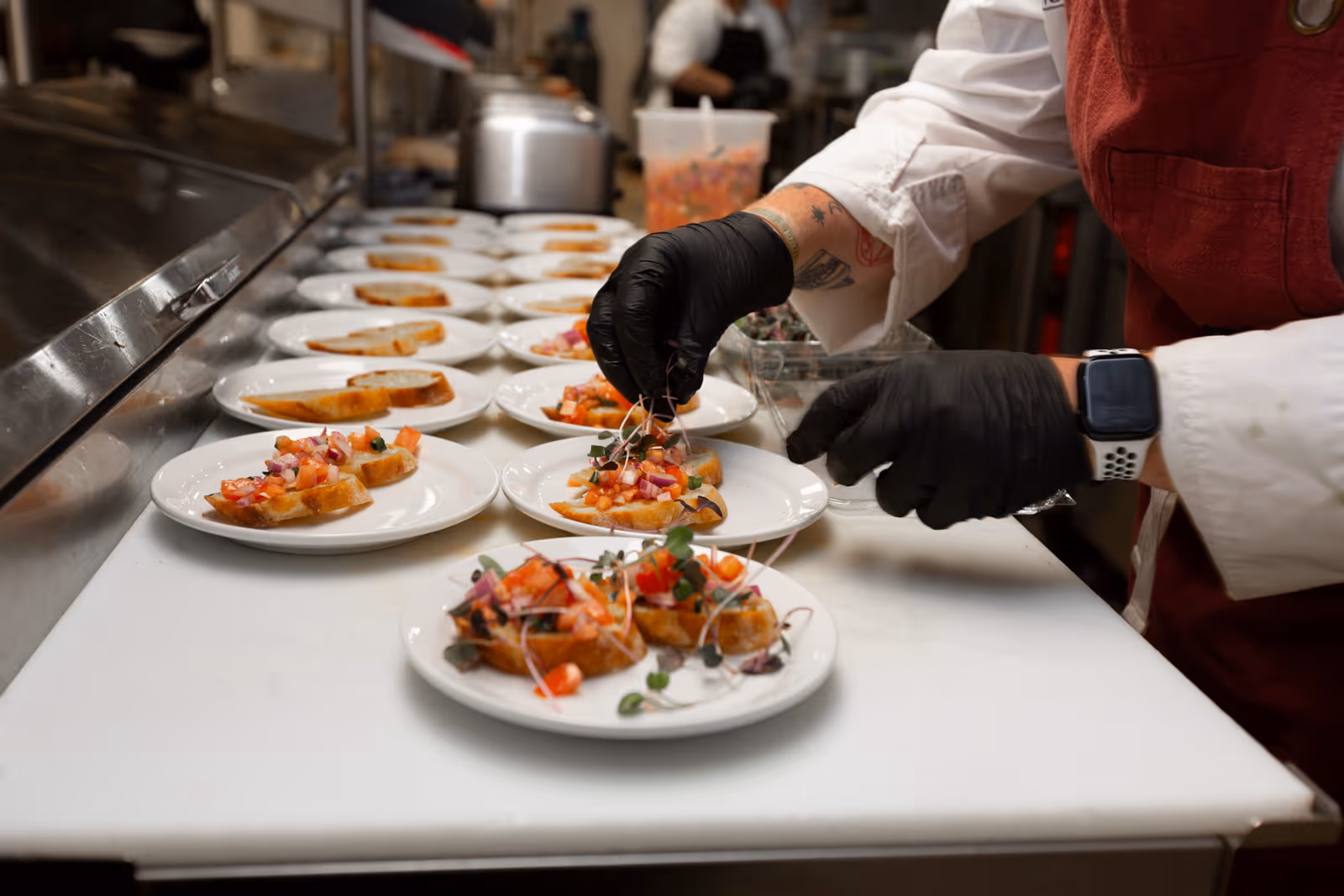 A chef wearing black gloves and a red apron is carefully placing microgreens on small appetizer plates with slices of toasted bread topped with diced tomatoes and onions, arranged in a kitchen setting.