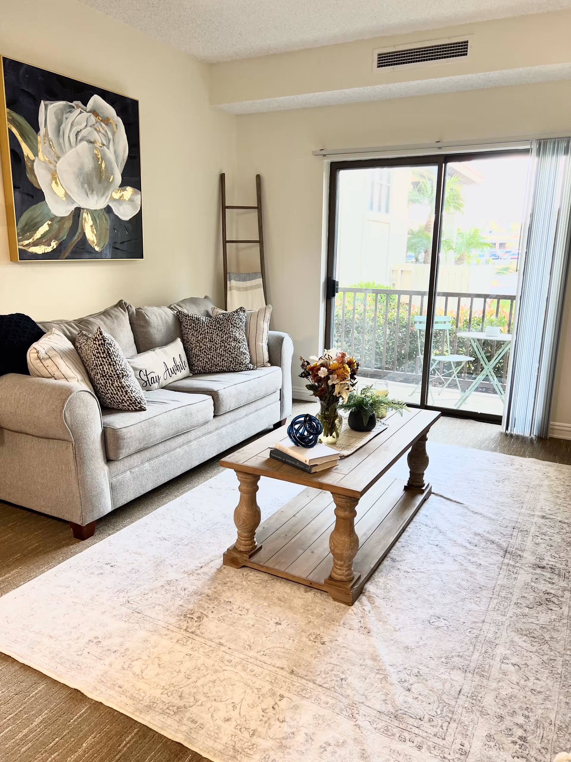 A cozy living room with a light gray sofa adorned with patterned and plain pillows, a wooden coffee table with decorative items and books, a large floral painting on the wall, a decorative ladder with a blanket, and a sliding glass door leading to a small outdoor patio with a table and chairs.