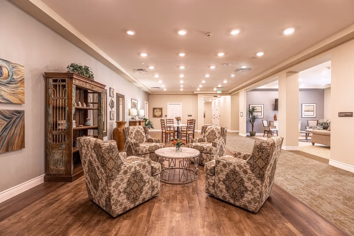 A spacious and well-lit common area in an assisted living facility featuring four patterned armchairs arranged around a round coffee table with a small flower vase. The room has wooden flooring, decorative wall art, a wooden bookshelf, and additional seating areas visible in the background with tables and chairs.