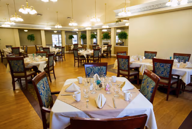 A spacious dining room with multiple round tables covered with white tablecloths and beige overlays, each set with folded napkins, cups, glasses, and silverware. The room has wooden floors, upholstered chairs with patterned fabric, hanging light fixtures, and large windows with potted plants on the windowsills.
