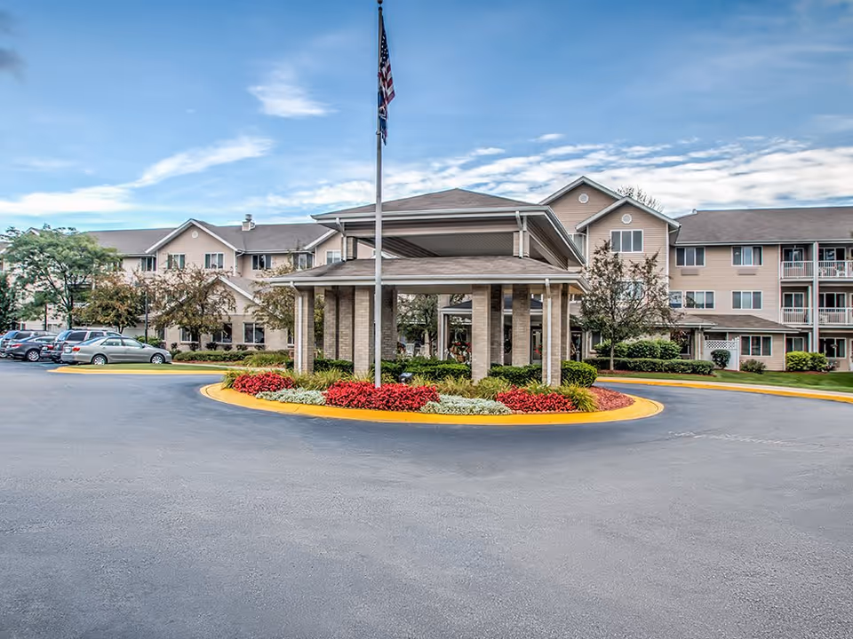 Exterior view of StoryPoint Wyoming senior living facility showing a circular driveway with a covered entrance, landscaped flower beds with red and white flowers, an American flag on a flagpole, and a multi-story beige building with balconies and windows under a partly cloudy sky.
