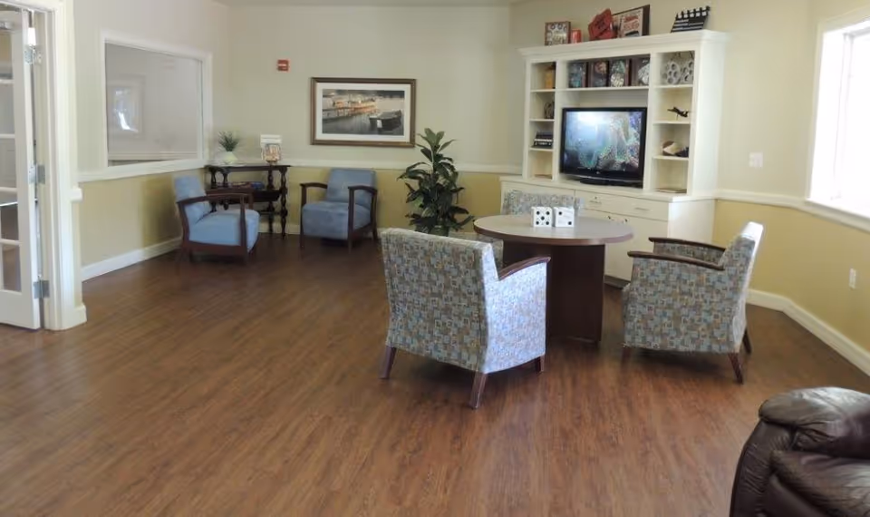 Spacious common living room with upholstered chairs around a round table, a white TV cabinet, framed art, and wood-look flooring.