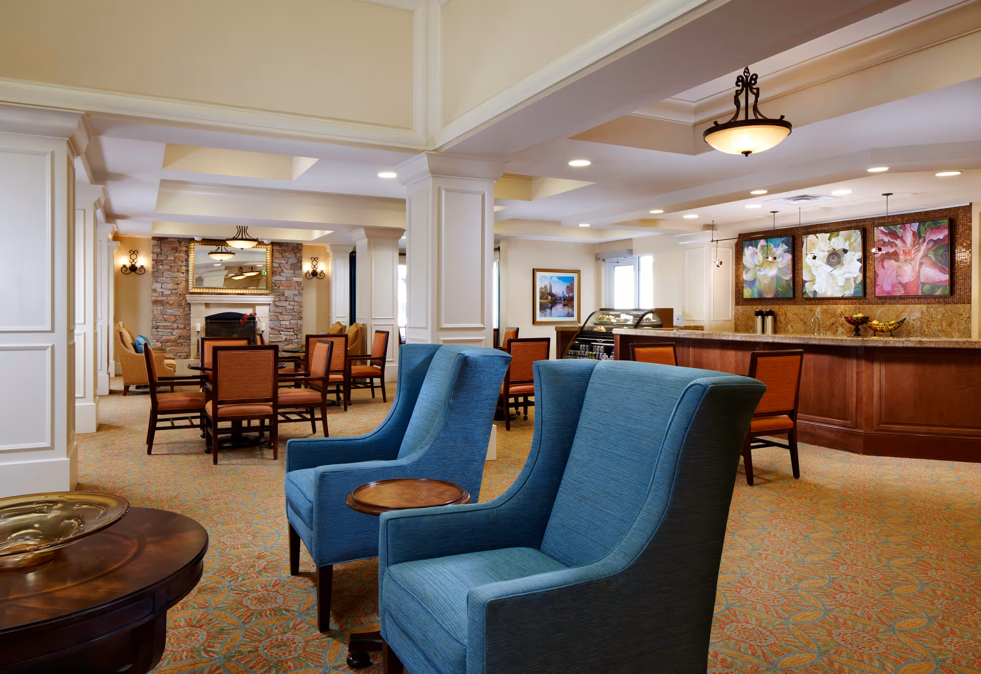 Bright senior living common area with blue lounge chairs, dining tables, a stone fireplace and a serving counter.