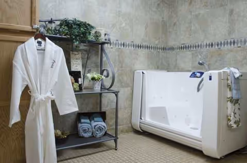 White bathrobe hangs near a walk-in tub with towels and plants in a tiled bathroom.