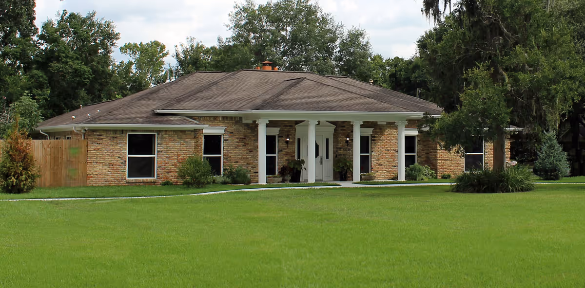 Single-story brick building with a dark shingled roof, white columns at the entrance, surrounded by green grass and trees in the background under a partly cloudy sky.