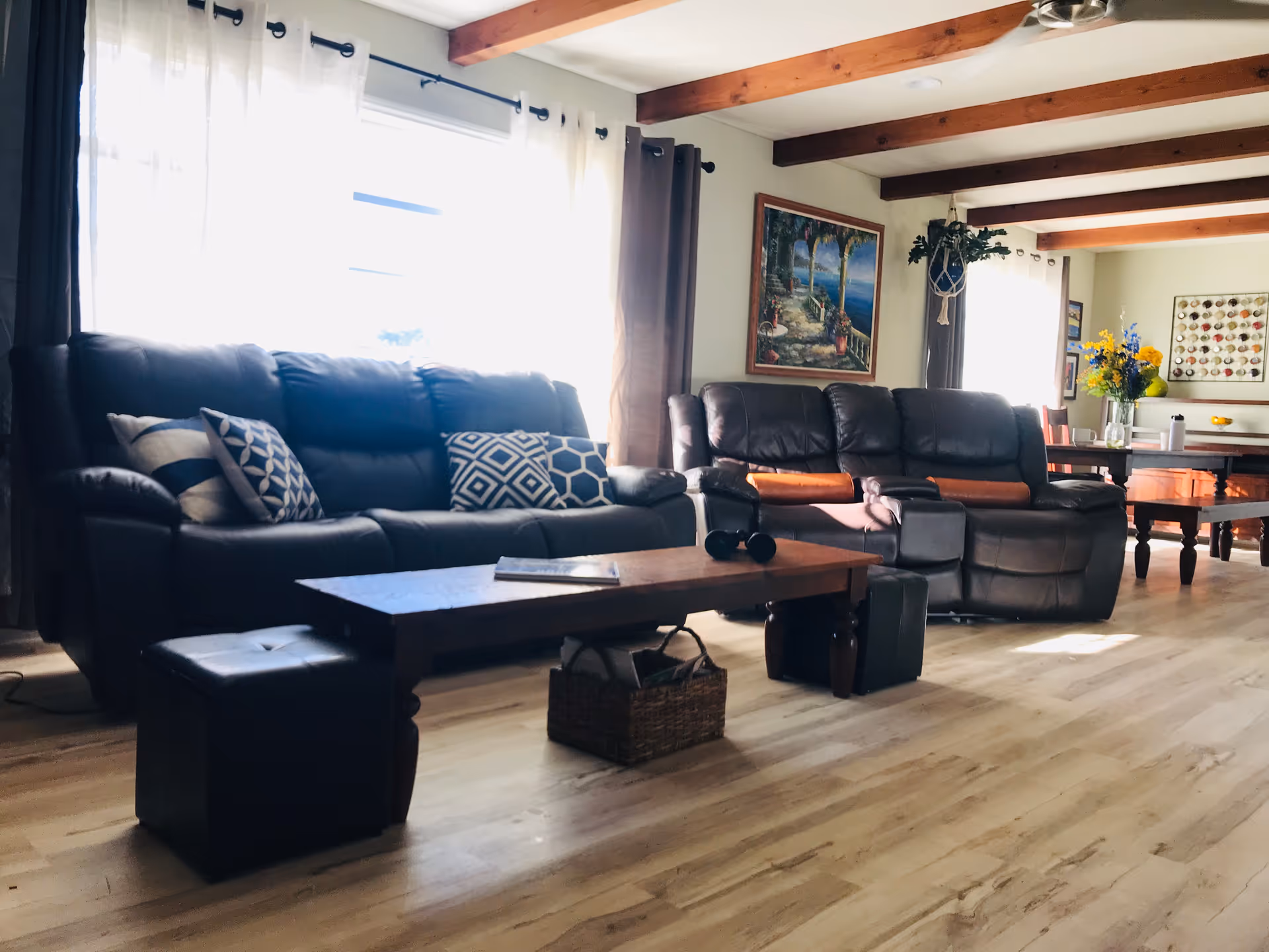 Bright living room with dark leather sofas, a wooden coffee table, exposed ceiling beams and a dining area visible in the background.