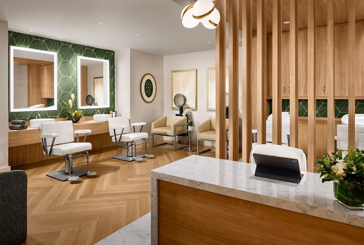 A modern salon area with two white styling chairs in front of a wooden counter with two illuminated mirrors on a green geometric patterned wall. There are two beige armchairs with a small table and decorative items in the background. The floor is wooden with a herringbone pattern, and a marble-topped reception desk with a tablet and a flower arrangement is in the foreground. Wooden vertical slats partially separate the space.