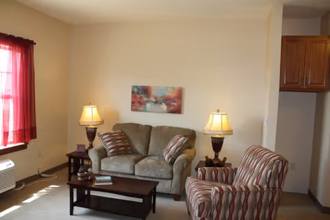 A cozy living room with a beige loveseat and a striped armchair. There are two side tables with matching lamps on either side of the loveseat. A wooden coffee table is in front of the loveseat with a book and decorative items on it. A colorful abstract painting hangs on the wall above the loveseat. A window with red curtains allows natural light into the room.