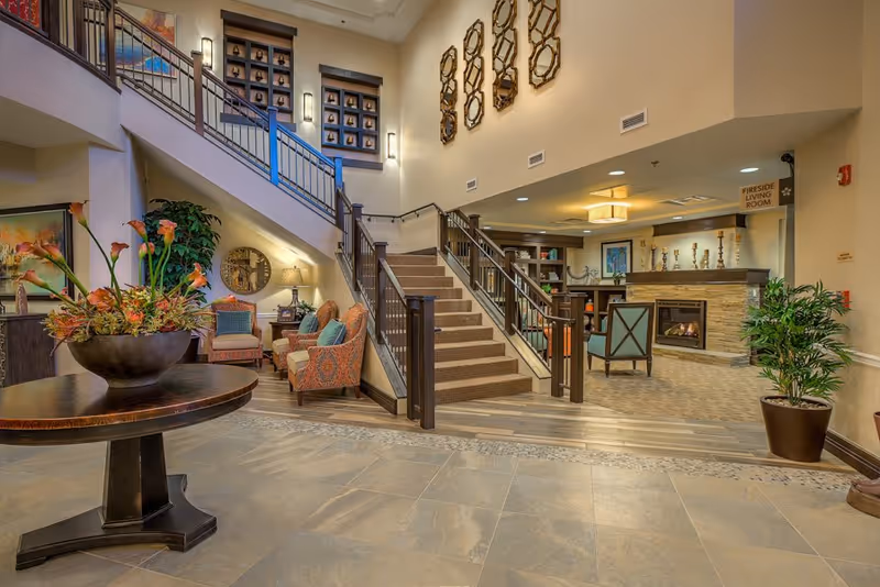Spacious, warmly lit assisted living lobby with a central staircase, seating areas, fireplace, and decorative plants.