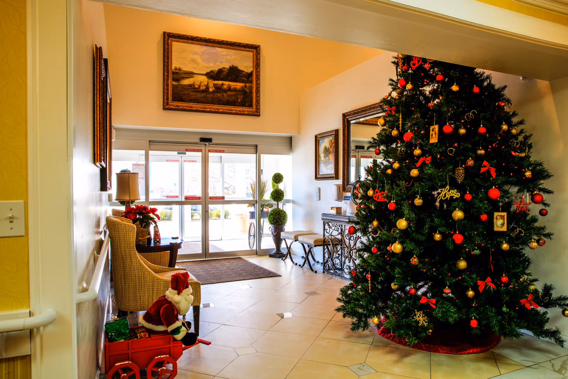 A senior-living lobby decorated for Christmas with a large ornamented tree, seating, and glass entry doors.