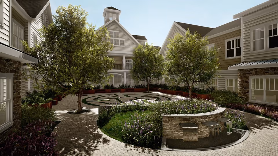 A peaceful outdoor courtyard at Broadview Senior Living at Purchase College featuring a circular garden with blooming flowers and several trees. The courtyard is surrounded by multi-story buildings with white siding and large windows, and there is a stone water feature in the foreground.