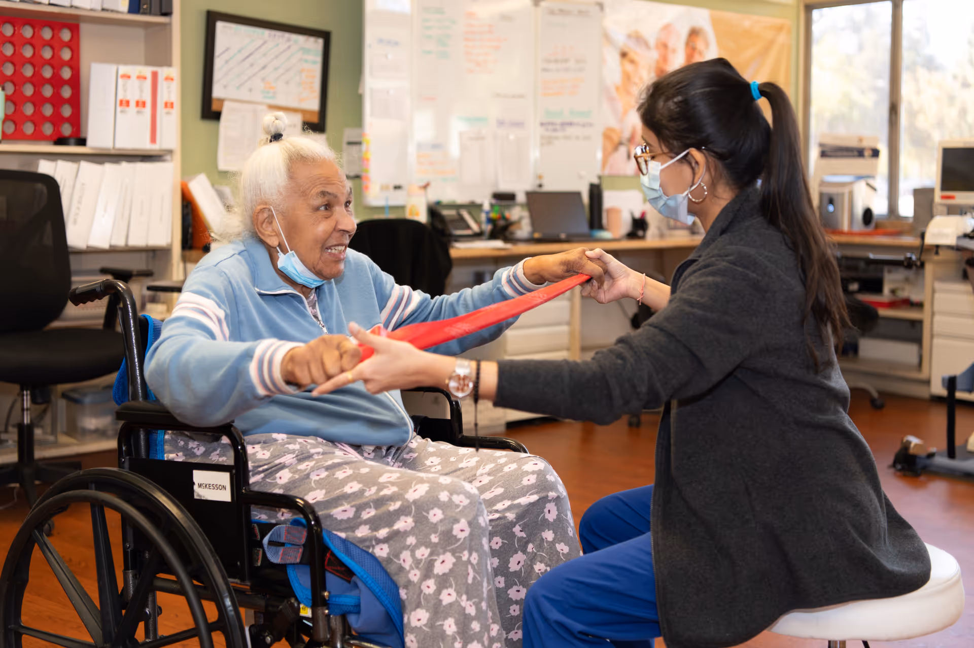 An elderly woman in a wheelchair is exercising with a physical therapist who is holding a red resistance band. Both are wearing face masks. The setting appears to be a rehabilitation or therapy room with office supplies and equipment in the background.