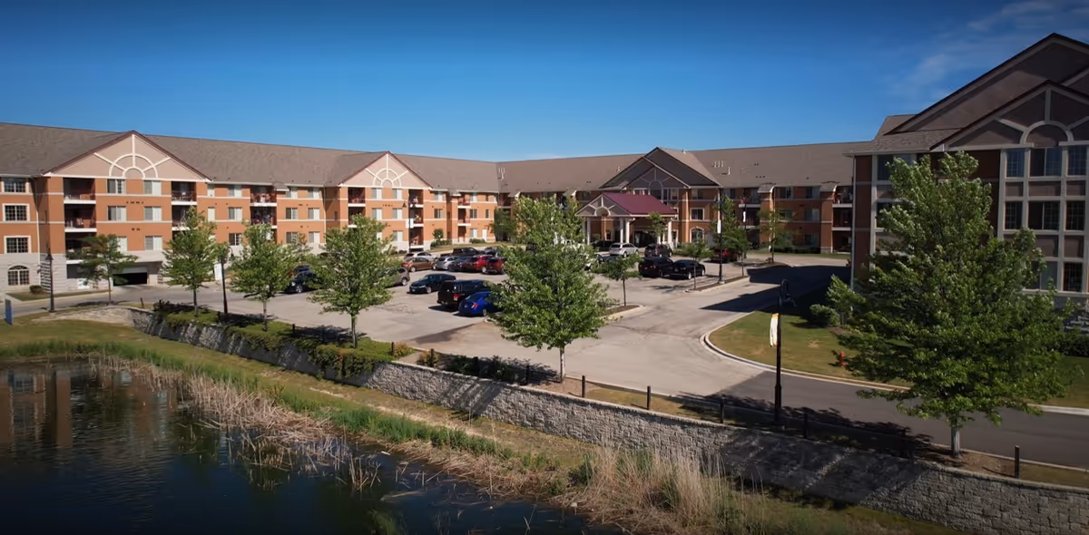 Exterior view of ProHealth Regency Senior Communities New Berlin showing a large multi-story building with balconies, a parking lot with several cars, landscaped trees, and a pond in the foreground under a clear blue sky.
