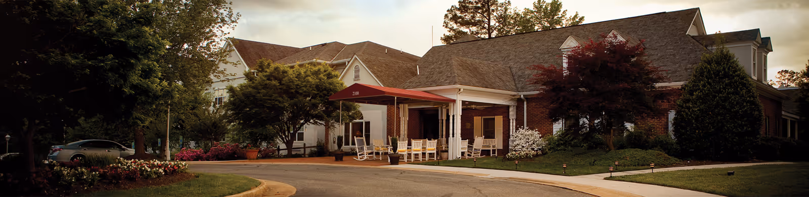 Exterior view of Brandermill Woods Healthcare Communities showing a single-story brick building with a covered entrance, white rocking chairs on the porch, surrounded by trees and landscaping under a cloudy sky.