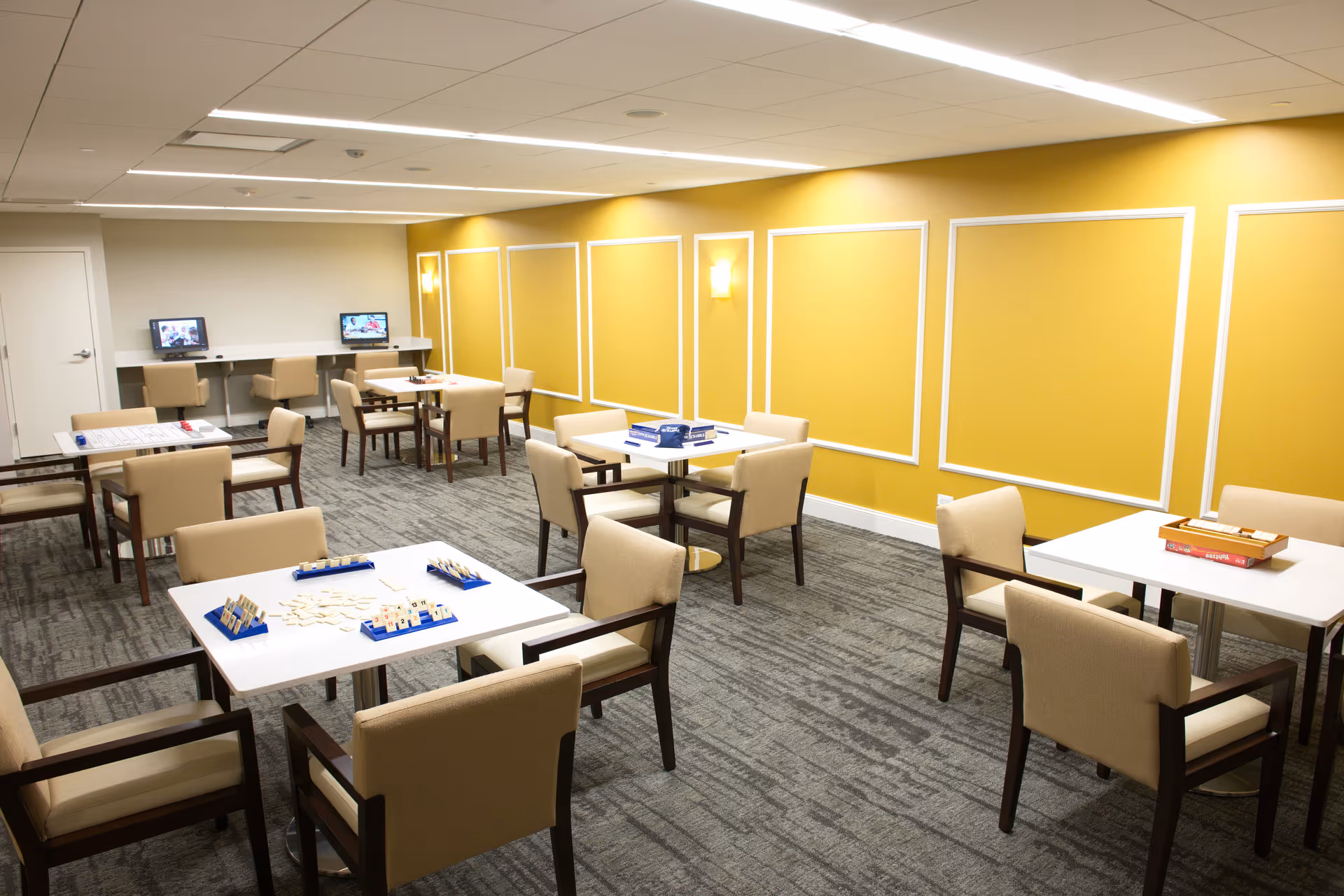 A well-lit activity room with several tables and beige cushioned chairs arranged for games. The tables have board games and game pieces set up. The walls are painted yellow with white trim accents, and there are two computer stations with chairs along one wall.