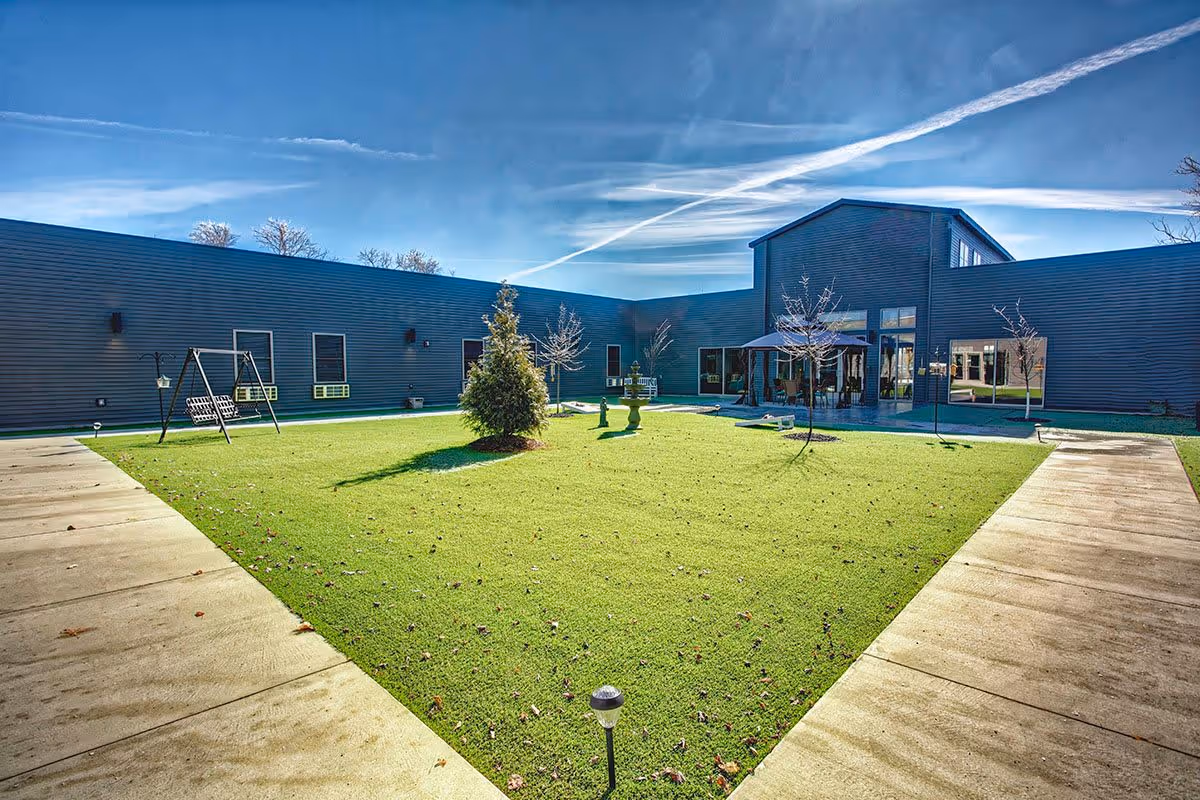 A courtyard area with green grass, small trees, a swing set, and a patio with outdoor seating under a canopy, surrounded by a modern building under a clear blue sky with visible contrails.