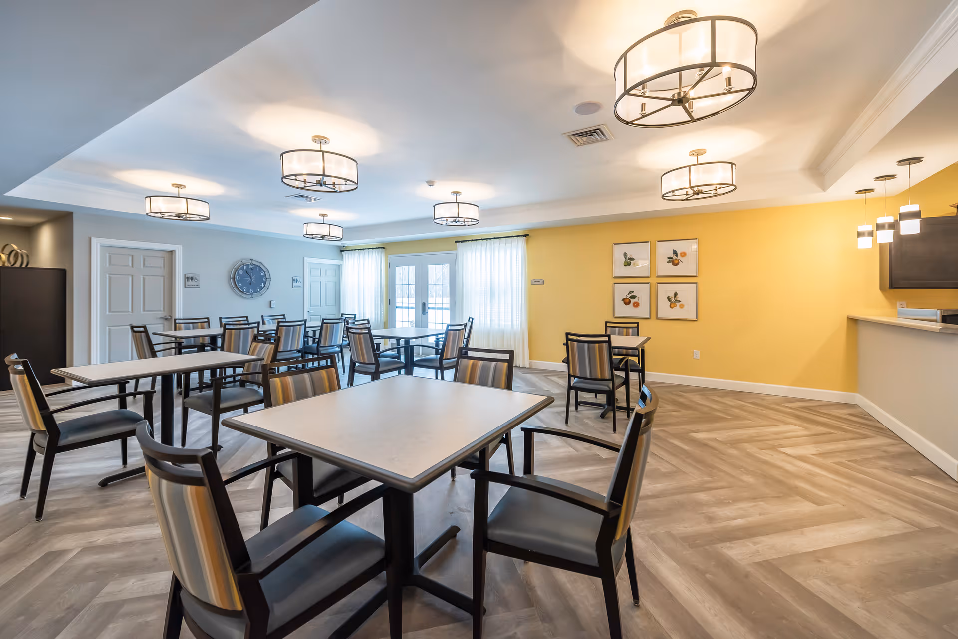 A bright dining room with multiple square tables and chairs arranged neatly. The room has a yellow accent wall with four framed fruit pictures, light gray walls, and large windows with sheer white curtains allowing natural light. Modern ceiling lights and pendant lights illuminate the space, and there is a counter area on the right side.