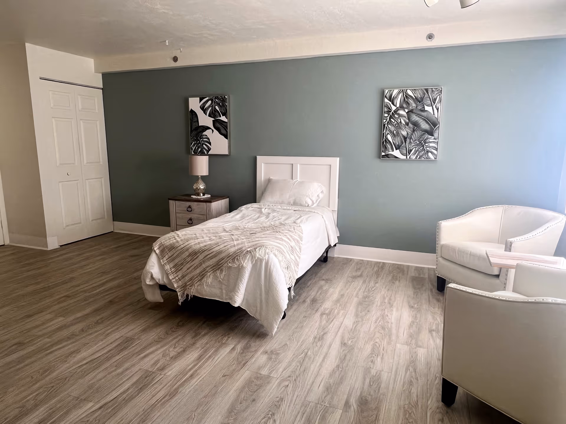 A simple bedroom with a single bed covered in white bedding and a beige throw blanket. There is a wooden nightstand with a lamp next to the bed. Two framed black and white botanical prints hang on a muted green accent wall behind the bed. The room has light wood flooring and two white armchairs with a small wooden table between them near a window.
