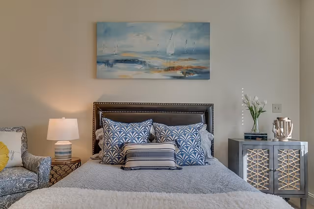 Bedroom with a made bed featuring patterned pillows and an upholstered headboard, flanked by a lamp and a decorative nightstand beneath wall art.