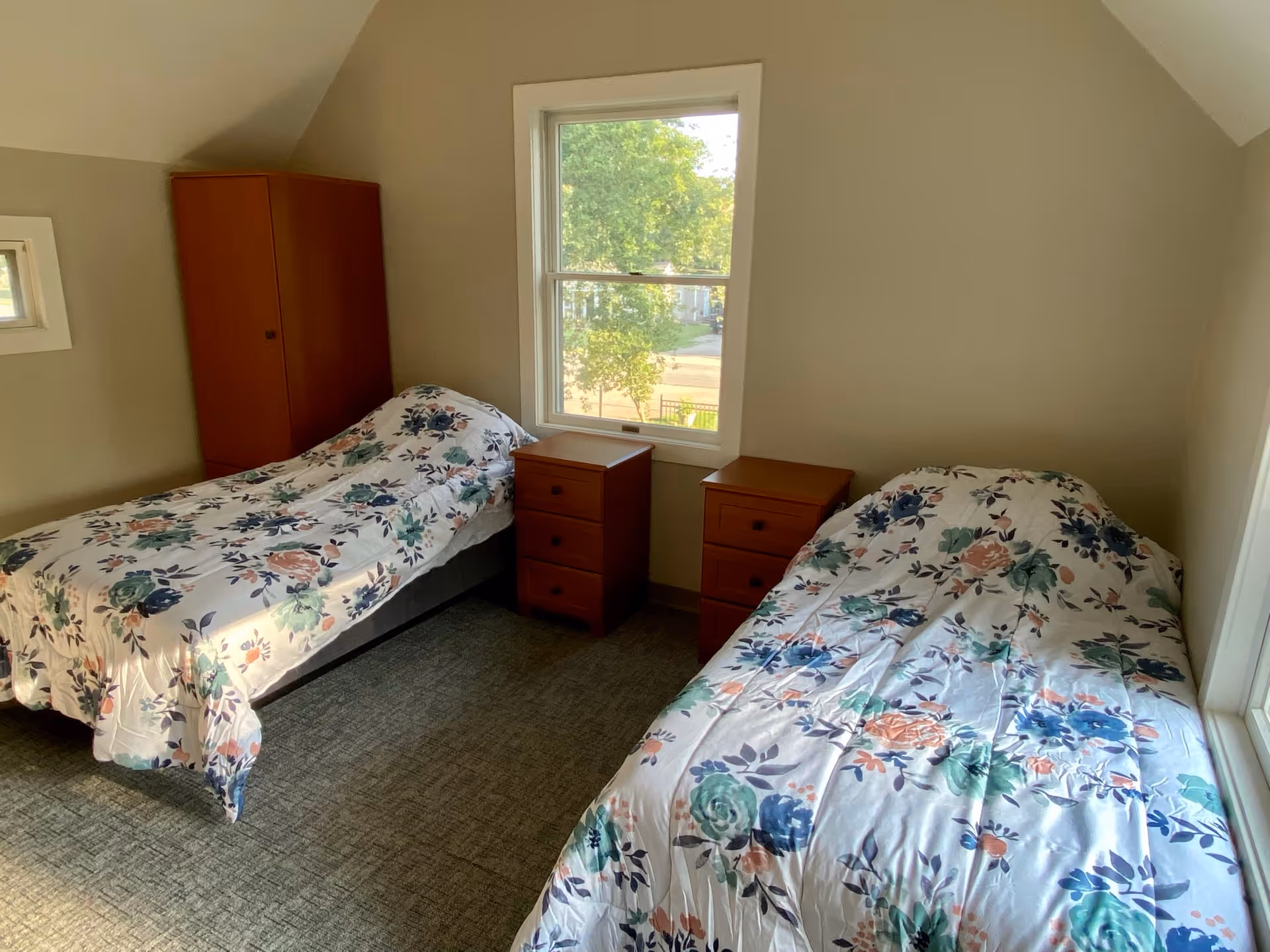 A bedroom with two single beds covered in floral patterned bedspreads. Between the beds are two wooden nightstands, and there is a wooden wardrobe in the corner. A window in the center of the wall lets in natural light and shows greenery outside.