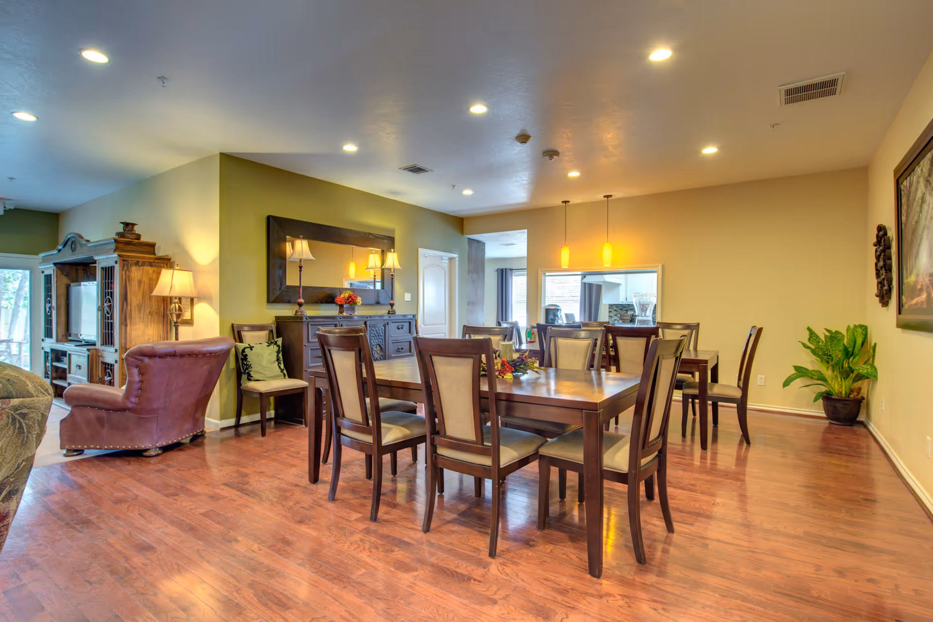 A spacious dining room with two wooden dining tables surrounded by chairs with beige cushions. The room has wooden flooring, a large mirror on the wall, two pendant lights hanging from the ceiling, and a potted plant in the corner. Adjacent to the dining area is a living space with a brown leather armchair, a wooden entertainment center, and a floor lamp.